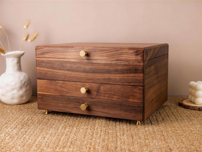 May include: A three-drawer wooden jewelry box with a rich brown finish. The box has gold-colored knobs and feet. The wood grain is visible. The box is sitting on a woven surface.