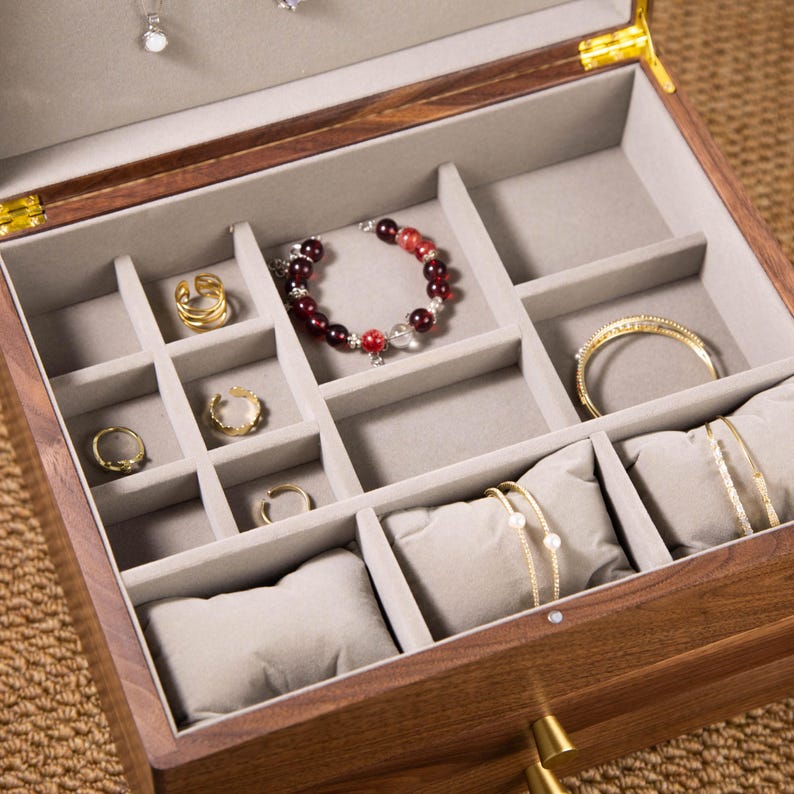 May include: Open wooden jewelry box with a gray velvet interior. The box contains various gold rings, bracelets, and a beaded bracelet with red and clear beads. The box is open, revealing multiple compartments.