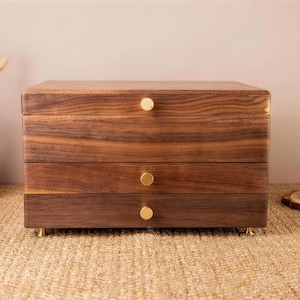 May include: A three-drawer wooden jewelry box with a rich, dark brown finish. The box has gold-colored knobs and feet. The wood grain is visible, adding to its natural aesthetic. The box is sitting on a woven surface.