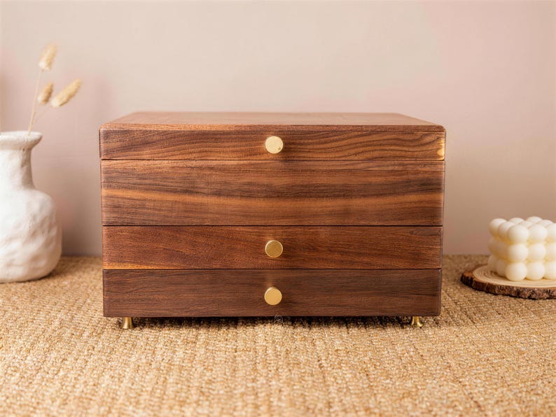 May include: A three-drawer wooden jewelry box with a rich, dark brown finish. The box has gold-colored knobs and feet. The wood grain is visible, adding to its natural aesthetic. The box is sitting on a woven surface.