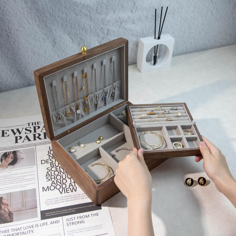May include: A wooden jewellery box with multiple compartments, lined with grey fabric. The box is open, revealing necklaces, rings, bracelets, and earrings. The box has a gold-coloured knob and is on a white surface.