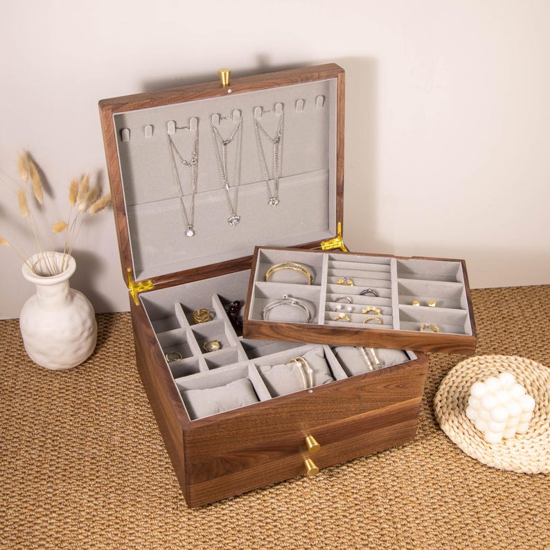 May include: A wooden jewelry box with a hinged lid and multiple compartments. The box is open, revealing necklaces, rings, bracelets, and other jewelry. The interior is lined with gray fabric. The box has a warm, brown finish and gold-colored hardware.
