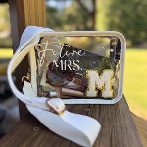 May include: A clear, rectangular crossbody bag with a white strap and gold hardware. The bag contains sunglasses, makeup, and a gold-trimmed letter "M" patch. The text "Future MRS." is written on the front of the bag.