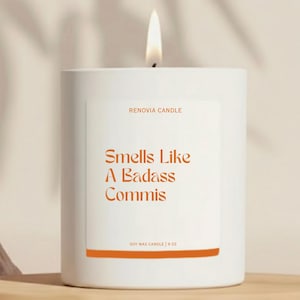 May include: A white soy wax candle with a lit wick. The label reads "Smells Like A Badass Commis" in orange text. The candle is 9 oz and is from Renovia Candle.