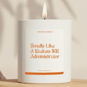 May include: A white candle with a lit wick. The label reads "Smells Like A Badass IRB Administrator" in orange text. The candle is labeled "RENOVIA CANDLE" and "SOY WAX CANDLE | 9 OZ".