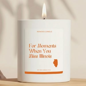 May include: A white soy wax candle with a lit wick. The label reads "For Moments When You Miss Illinois" with an orange outline of the state. The candle is 9 oz.