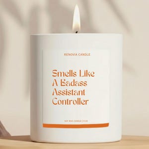 May include: A white soy wax candle with a lit wick. The label reads "Smells Like A Badass Assistant Controller" in orange text. The candle is 9 oz and from Renovia Candle.
