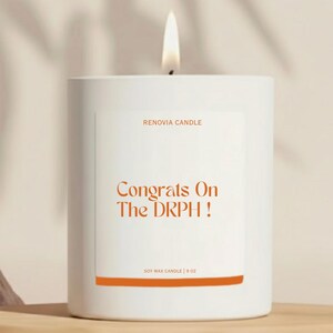 May include: A white soy wax candle with a lit wick. The label reads "Congrats On The DRPH!" in orange text, with "RENOVIA CANDLE" above and "SOY WAX CANDLE | 9 OZ" below. The candle sits on a wooden shelf.