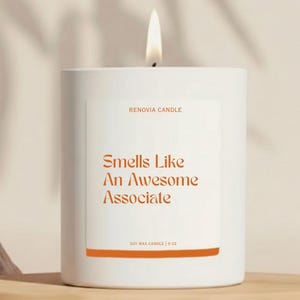 May include: A white soy wax candle with a lit wick. The label reads "Smells Like An Awesome Associate" in orange text. The candle is 9 oz.