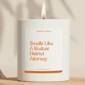 May include: A white soy wax candle with a lit wick. The label features the text "Smells Like A Badass District Attorney" in orange. The candle is 9 oz.
