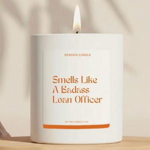May include: A white soy wax candle with a lit wick. The label reads "Smells Like A Badass Loan Officer" in orange text. The candle is 9 oz and from Renovia Candle.