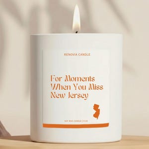 May include: A white candle with a lit wick. The label reads "For Moments When You Miss New Jersey" with a state outline. The candle is from Renovia Candle and is a soy wax candle, 9 oz.