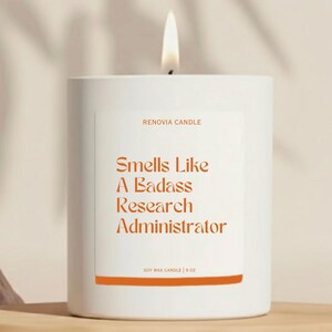 May include: A white soy wax candle with a lit wick. The candle has a label with the text "Smells Like A Badass Research Administrator" in orange. The candle is 9 oz.