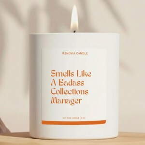 May include: A white soy wax candle with a lit wick. The label reads "Smells Like A Badass Collections Manager" in orange text. The candle is 9 oz and from Renovia Candle.