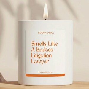 May include: A white candle with a lit wick. The label reads "Smells Like A Badass Litigation Lawyer" in orange text. The candle is labeled "RENOVIA CANDLE" and "SOY WAX CANDLE | 9 OZ".