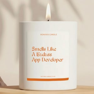 May include: A white soy wax candle with a lit wick. The label reads "Smells Like A Badass App Developer" in orange text. The candle is 9 oz and from Renovia Candle.