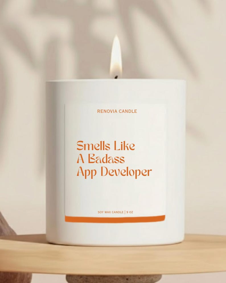 Op de afbeelding: Een witte kaars met een brandende lont. Het etiket zegt "Smells Like A Badass App Developer" in oranje tekst. De kaars is 255g en van Renovia Candle.