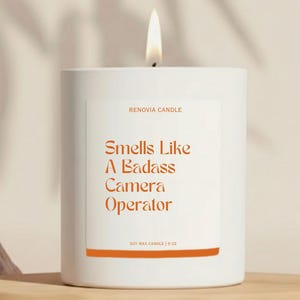 May include: A white soy wax candle with a lit wick. The label reads "Smells Like A Badass Camera Operator" in orange text. The candle is 9 oz and sits on a wooden shelf.