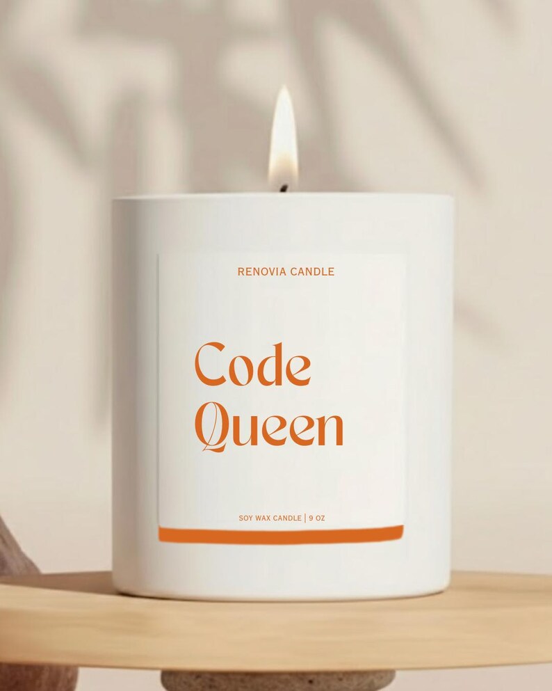 Op de afbeelding: Een brandende witte kaars met de tekst "Code Queen" in oranje. De kaars zit in een witte cilindrische houder met een label met de tekst "RENOVIA CANDLE" en "SOY WAX CANDLE | 9 OZ". De kaars staat op een houten plank.