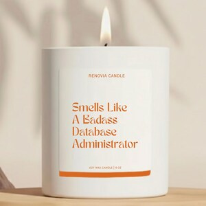 May include: A white Renovia Candle with a lit wick. The label reads "Smells Like A Badass Database Administrator" in orange text. The candle is soy wax and weighs 9 oz.