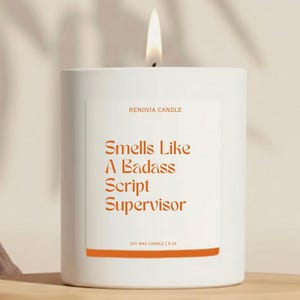 May include: A white candle with the text "Smells Like A Badass Script Supervisor" in orange. The candle is labeled "RENOVIA CANDLE" and "SOY WAX CANDLE | 9 OZ". A lit flame sits atop the candle.