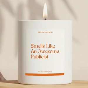 May include: A white soy wax candle with a lit wick. The label reads "RENOVIA CANDLE" and "Smells Like An Awesome Publicist." The candle is 9 oz. and sits on a wooden shelf.