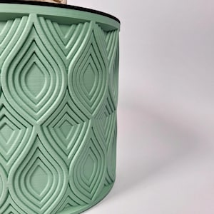 Peut inclure: Un récipient cylindrique vert clair avec un motif en relief répété de formes en goutte d'eau. Le design est symétrique et crée une surface texturée. Le bord supérieur est noir.