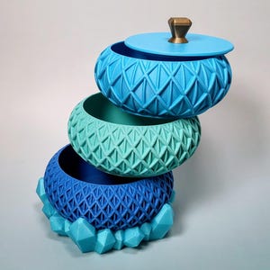 May include: Three-tiered decorative storage bowls in shades of blue and teal. The top bowl has a lid with a gold-colored knob. Each bowl features a geometric diamond pattern. The bottom bowl rests on a base of blue crystal-shaped accents.