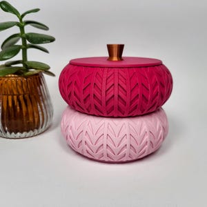 Peut inclure: Deux boîtes décoratives rondes à couvercle, empilées l'une sur l'autre, dans des tons de rose et de rouge. Les boîtes présentent un motif texturé en forme de chevrons. Une petite plante succulente dans un récipient en verre se trouve à gauche.