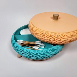 Peut inclure: Un récipient rond bicolore avec un couvercle, conçu pour ranger de petits objets. La base est turquoise avec un motif de vagues sculptées, et le couvercle est de couleur pêche avec un design similaire. Le récipient contient des cotons-tiges et des disques.