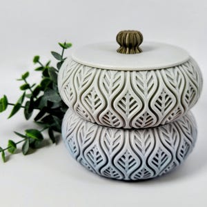 Może przedstawiać: Dwuczęściowy dekoracyjny pojemnik z pokrywką. Pojemnik jest biały ze wzorem liści i gałką w kolorze brązu. Dolna część ma gradient niebiesko-biały. Pojemnik stoi na białej powierzchni z zielonymi liśćmi.