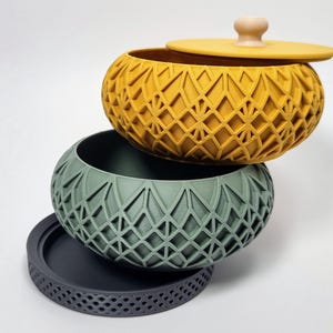 Peut inclure: Ensemble de trois bols décoratifs. L'ensemble comprend un bol jaune moutarde avec un couvercle, un bol vert sauge et une base gris foncé. Chaque pièce présente un motif géométrique en treillis. Le couvercle a un petit bouton en bois.