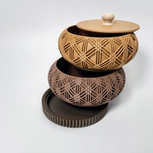 Peut inclure: Ensemble de trois bols décoratifs bruns. Les bols présentent un motif géométrique en treillis et un couvercle avec un petit bouton. La pièce inférieure est un plateau rond avec un bord cannelé. Les bols sont empilés les uns sur les autres.