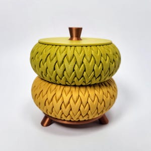 Peut inclure: Un récipient décoratif en deux parties avec un motif tissé texturé. Le dessus est vert olive et le dessous est jaune doré. Il a une tige et des pieds couleur cuivre, et un couvercle assorti avec un bouton en cuivre.