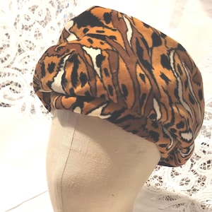 May include: A tiger print hat, wooden bracelet, and earrings are displayed on a mannequin head. The hat features a brown, black, and white pattern. The bracelet and earrings are made of wood and have a natural finish.