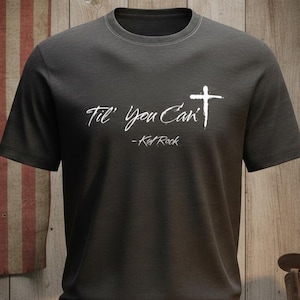 Puede incluir: Camiseta gris oscuro con la frase "Til' You Can't" y una cruz, atribuida a Kid Rock. La camiseta se muestra sobre un fondo de una bandera americana desgastada y tablones de madera.