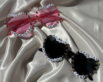 May include: Two pairs of heart-shaped sunglasses. One pair has pink lenses and the other has black lenses. Both pairs have white beaded text that reads "Olivia Rodrigo Tour Unraveled". The frames are decorated with gold stars.