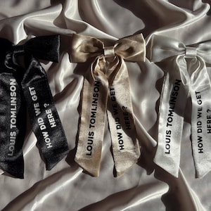 May include: Three satin hair bows in black, champagne, and white. Each bow has long streamers with the text "LOUIS TOMLINSON" and "HOW DID WE GET HERE ?" printed in a contrasting colour. The bows are arranged on a white satin surface.
