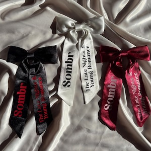 Könnte beinhalten: Drei Satin-Haarschleifen in Schwarz, Weiß und Burgunderrot. Jede Schleife hat lange Bänder mit dem Wort "Sombr" und dem Satz "The Late Nights & Young Romance" aufgedruckt. Die Schleifen sind auf einem seidigen, cremefarbenen Stoff angeordnet.