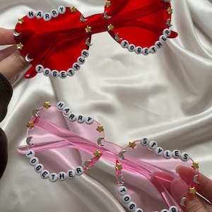 May include: Two pairs of heart-shaped sunglasses. One pair is red with the words "HARRY STYLES TOGETHER" spelled out in white beads. The other pair is pink with the same text. Both have gold star and moon accents.