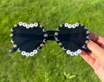 Könnte beinhalten: Herzförmige Sonnenbrille mit dunklen Gläsern, verziert mit kleinen goldfarbenen Sternen und silberfarbenen Mondsicheln am Rand. Die Rahmen sind mit weißen Buchstabenperlen versehen, die links OASIS TOUR und rechts LOVE 2027 buchstabieren.