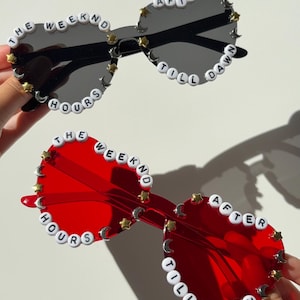 May include: Two pairs of heart-shaped sunglasses, one with black lenses and one with red lenses. Both frames are decorated with white letter beads spelling THE WEEKND AFTER HOURS TILL DAWN, interspersed with small gold star and silver moon charms.