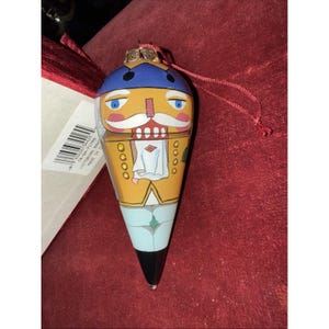 Może przedstawiać: Ozdoba świąteczna w kształcie dziadka do orzechów. Ozdoba ma niebieski kapelusz, białe wąsy i żółtą kurtkę. Dolna połowa jest jasnoniebieska i czarna. Do zawieszenia przymocowany jest czerwony sznurek.