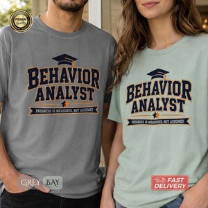 Könnte beinhalten: Ein graues T-Shirt und ein hellgrünes T-Shirt, beide mit einer dunkelblauen Grafik, die eine Doktorhut-Ikone, den Text BEHAVIOR ANALYST, ein kleines Puzzleteil und den Bannertext PROGRESS IS MEASURED, NOT ASSUMED auf der Vorderseite zeigt.