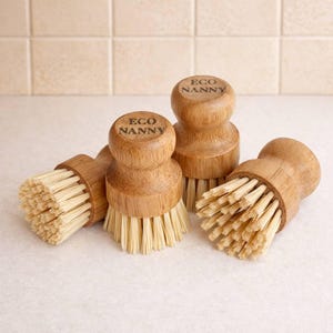 May include: Four wooden dish brushes with natural bristles. Each brush has a round wooden handle with the text "ECO NANNY" printed on top. The bristles are a light beige color, and the wood has a light brown tone.