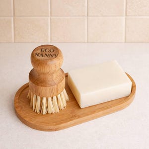 Puede incluir: Un juego de limpieza de cocina de bambú que incluye un cepillo para platos con el texto "ECO NANNY", una pastilla de jabón rectangular y una bandeja ovalada. El cepillo tiene cerdas claras. El jabón es de color crema claro.