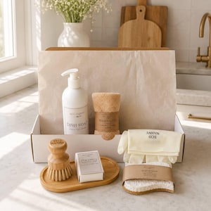 May include: A white box containing dish soap, a washcloth, a dish brush, a bar of coconut dish soap, dish gloves, and a dish sponge. The products are arranged on a countertop with a vase of flowers in the background. The dish soap bottle has a pump.