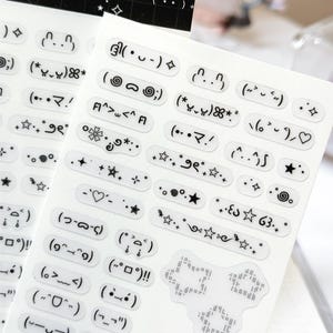 May include: A sheet of clear stickers featuring black printed kaomoji emoticons, stars, hearts, and decorative symbols. The top header text reads INGSOUF STUDIO [ no.2 - kaomoji ] with a small emoticon. The stickers are arranged in rows.