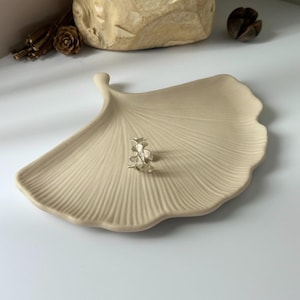 Peut inclure: Un plat en céramique beige en forme de feuille avec une surface texturée. Une petite bague florale argentée repose sur le plat. Le plat est sur une surface blanche, avec une sculpture en bois et d'autres éléments décoratifs en arrière-plan.