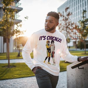 May include: White long-sleeve sweatshirt with the text "IT'S CHICAGO!" in purple and a graphic of two football players. The sweatshirt is worn by a person standing outdoors near a building and a staircase.